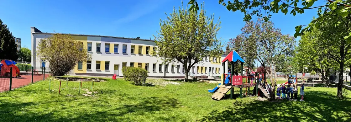 Budynek przedszkola od strony ogrodu: parterowo-piętrowy obiekt z jasną elewacją, przed nim trawnik i plac zabaw z kolorowymi urządzeniami.