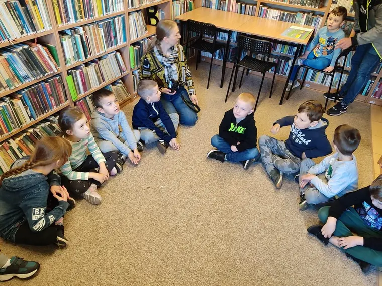 Dzieci siedzą na podłodze wśród regałów z książkami podczas wizyty w bibliotece.